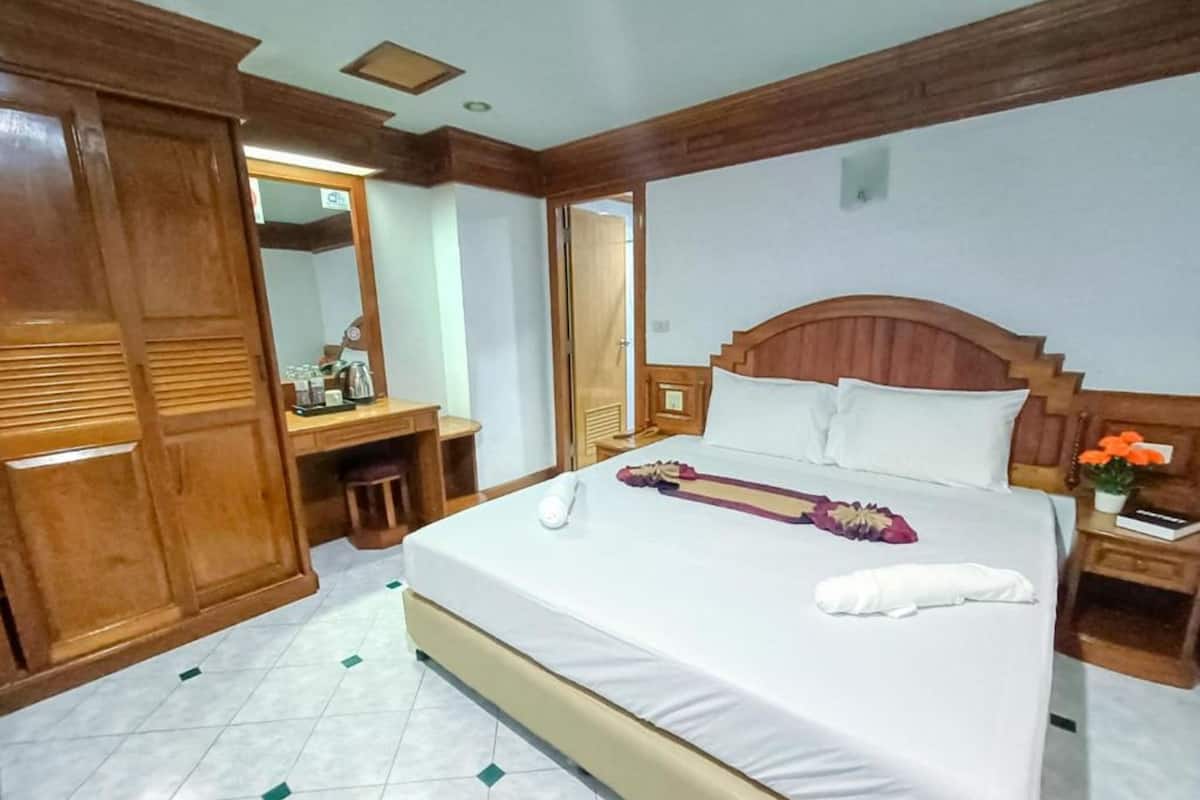 A hotel room with a large bed, wooden wardrobe, a desk with a chair, and a nightstand with a lamp.