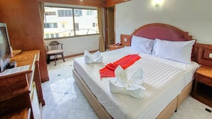 A hotel room with a large bed, a TV, a desk, and a window with a view of buildings. - Seascape (Patong)