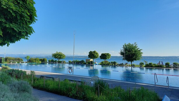 A swimming pool with clear blue water, surrounded by greenery and trees, with a view of the sea and mountains in the background.