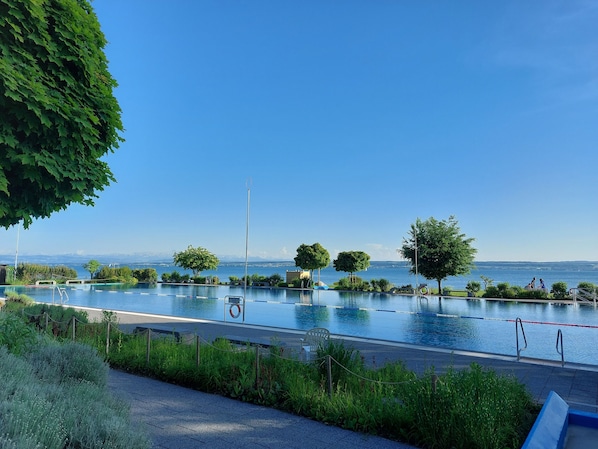 Ein Schwimmbecken mit klarem, blauem Wasser, umgeben von Grünpflanzen und Bäumen, mit Blick auf das Meer und die Berge im Hintergrund.