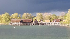 A row of wooden cabins with thatched roofs on stilts over a calm lake, surrounded by trees.