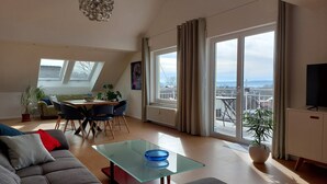 A modern living room with a glass coffee table, grey sofa, dining area with chairs, and large windows offering a city view.