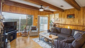 A spacious living room with wooden walls, a ceiling fan, a large TV, a sofa, a chair, and a dining table.