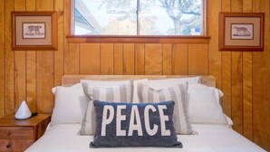 A bedroom with wooden paneling, a bed with white bedding, a pillow with the word "PEACE," and framed animal pictures on the walls.