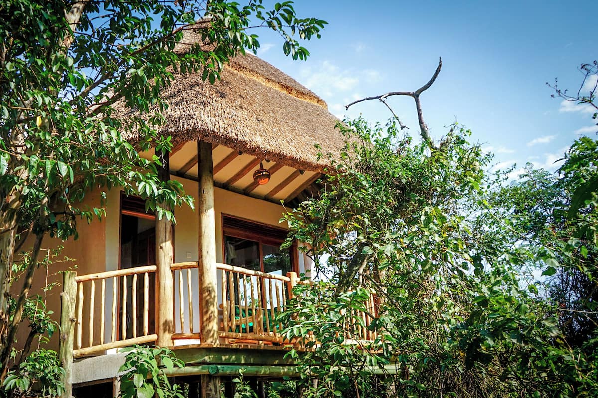 A thatched-roof bungalow with a wooden balcony, surrounded by lush greenery.