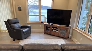 A living room with a black leather recliner, a flat-screen TV on a wooden stand, and a view of a lake through large windows. - Newer Construction waterfront house with AC and WiFi in Barnstead (Barnstead)