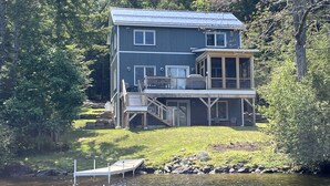 A two-story house with a deck and a dock extending into a lake, surrounded by trees. - Newer Construction waterfront house with AC and WiFi in Barnstead (Barnstead)