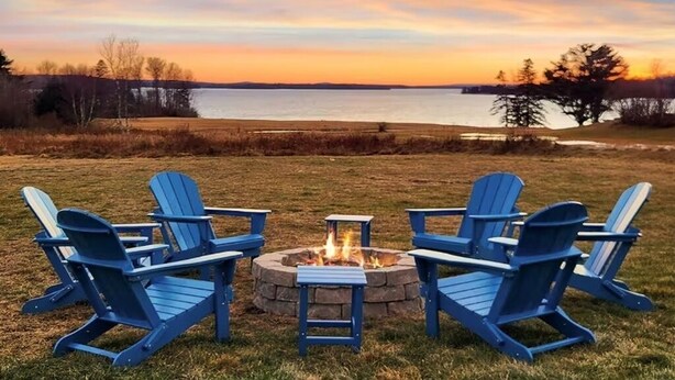 A fire pit with four blue Adirondack chairs arranged around it, set against a backdrop of a lake and trees during sunset. - 5 Min to Lamoine State Park & Lamoine Beach (Lamoine)