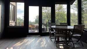 A room with a large table and chairs, large windows, and a view of trees and a deck outside.