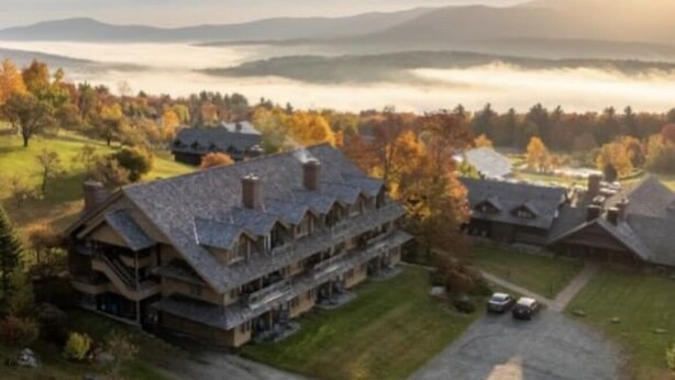 A large, multi-story lodge with a prominent central building and several smaller structures, surrounded by a lush landscape and a distant mountain range.