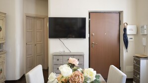 A hotel room with a flat-screen TV mounted on the wall, a wooden door, a small cabinet with drawers, and a floral arrangement on the table. - Charme & Chic Studio (Salerno)