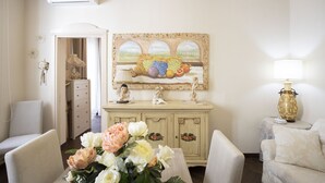 A room with a white sofa, a dining table with a vase of flowers, a cream-colored cabinet, and a wall-mounted painting. - Charme & Chic Studio (Salerno)