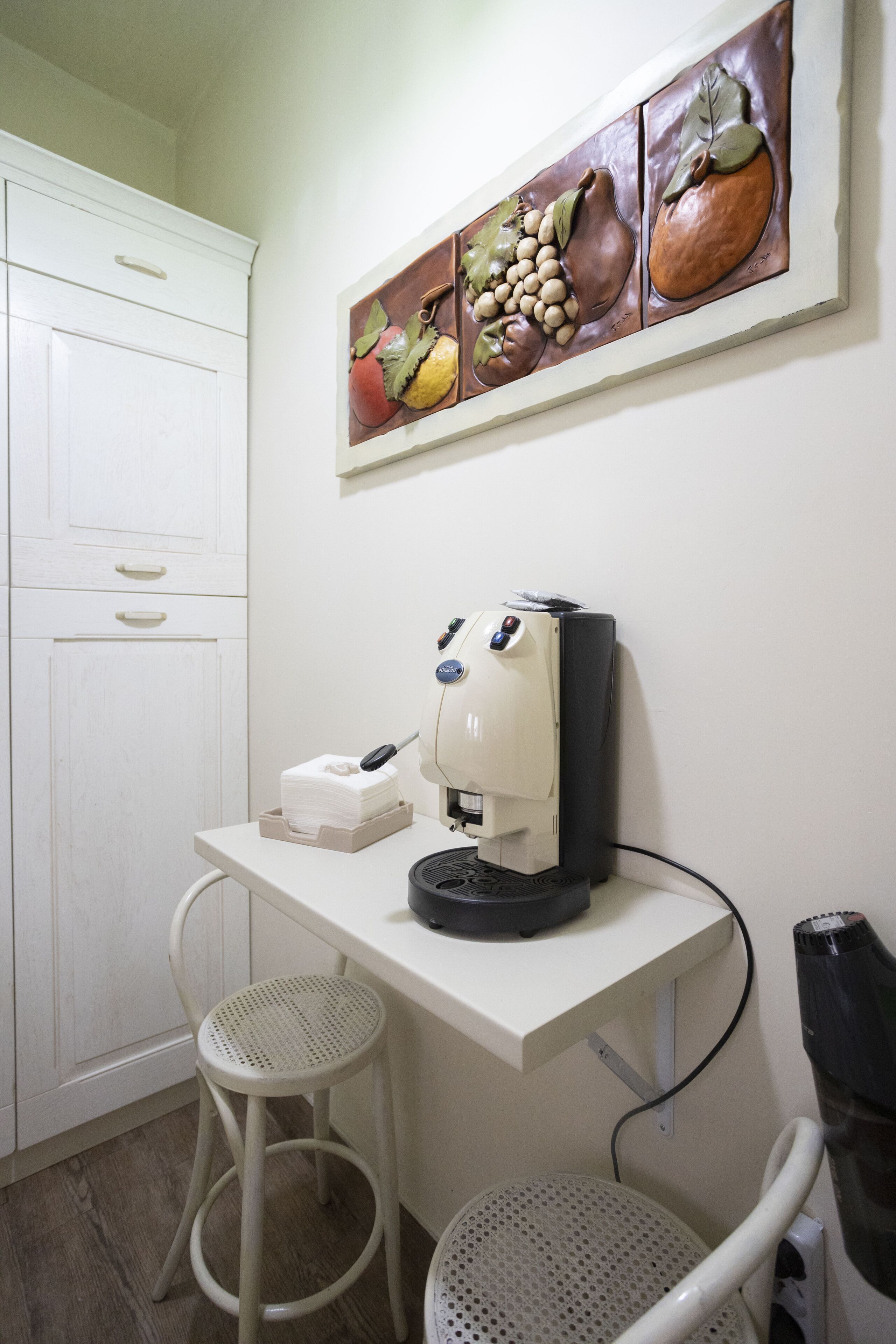 Una kitchenette con cafetera, un armario blanco y una obra de arte con motivos de frutas en la pared.