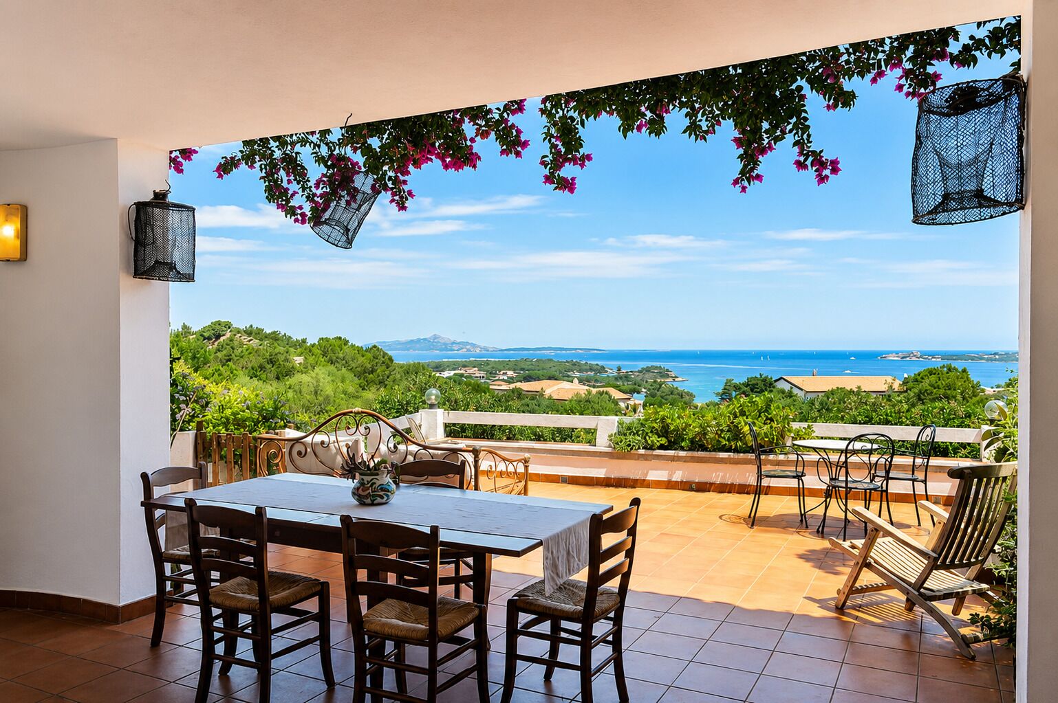 A terrace with a table and chairs, overlooking a coastal landscape with greenery and the sea.