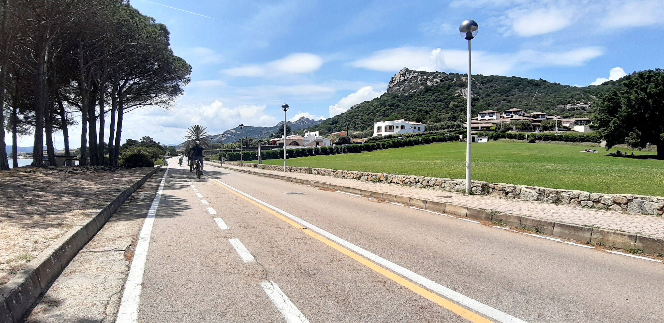A paved road with a sidewalk, trees, and a grassy area with a mountain in the background.