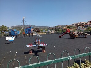 A playground with various play equipment, including swings and a slide, set against a backdrop of buildings and hills.