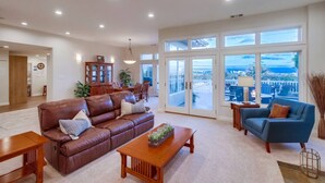 A spacious living room with a brown leather sofa, a blue armchair, and a wooden coffee table. There is a dining area with a wooden cabinet and chairs. Large windows offer a view of the sea and a balcony.
