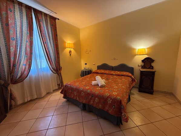 A hotel room with a bed, two wall lamps, a clock, and a window with curtains. - Villa le Camelie Siena (Siena)