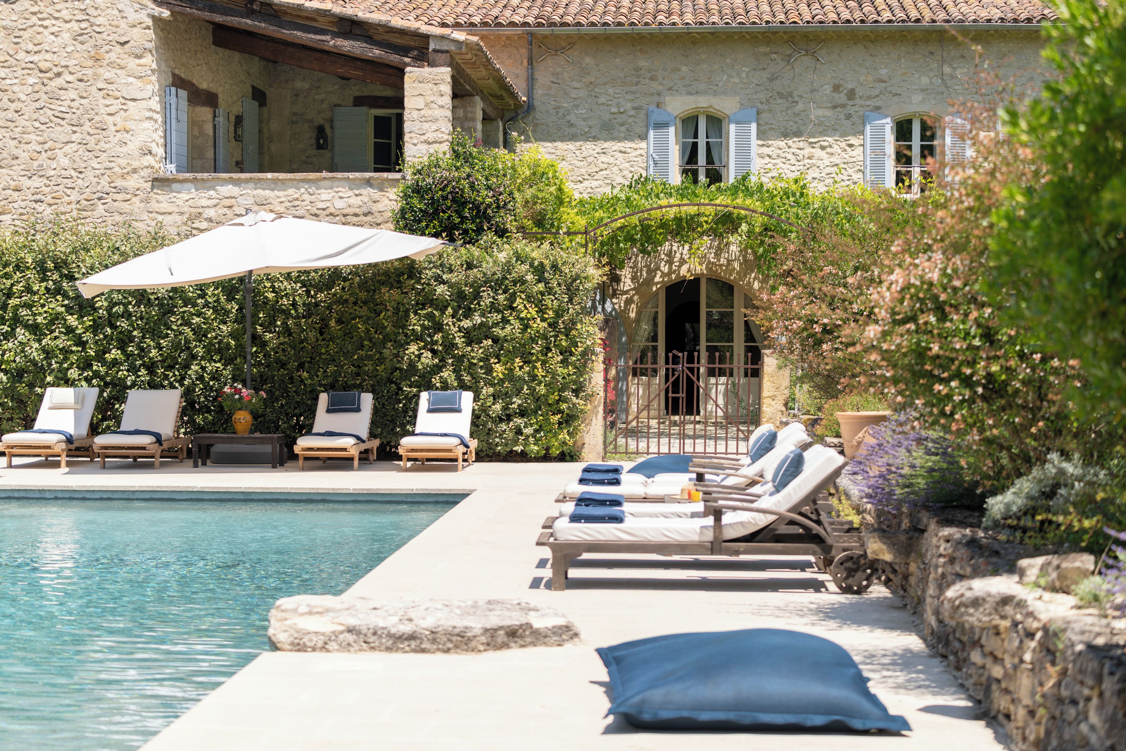 A pool area with lounge chairs and umbrellas, surrounded by a stone building with shutters.