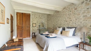Una camera da letto con parete in pietra, porta in legno, un letto con cuscini e coperta, un comodino con lampada e uno sgabello.