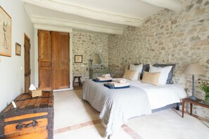 Ein Schlafzimmer mit Steinwand, Holztür, Bett mit Kissen und Decke, Nachttisch mit Lampe und einem kleinen Hocker.