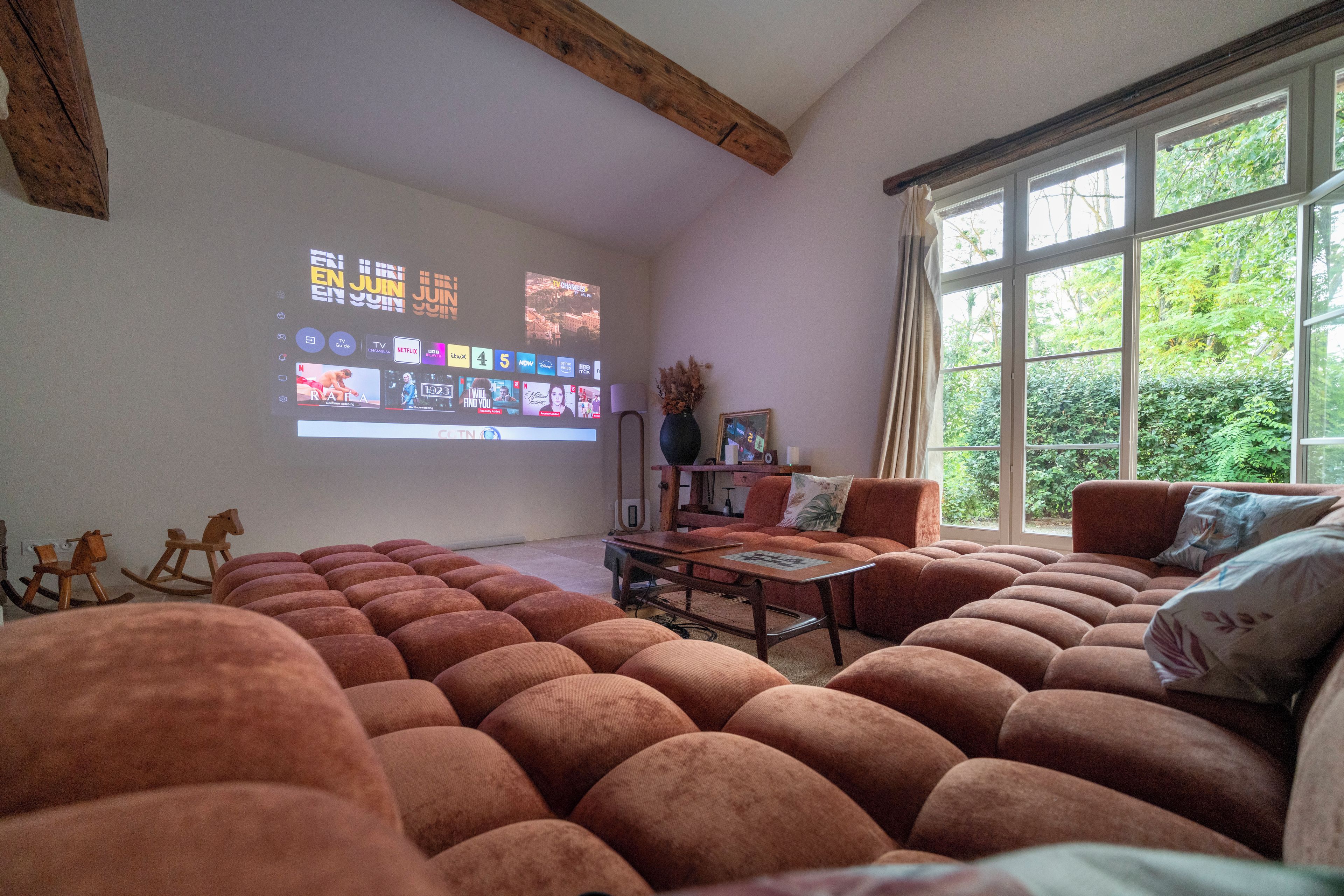 A living room with a large screen displaying a TV guide, a sofa set, a coffee table, and a window with a view of greenery.