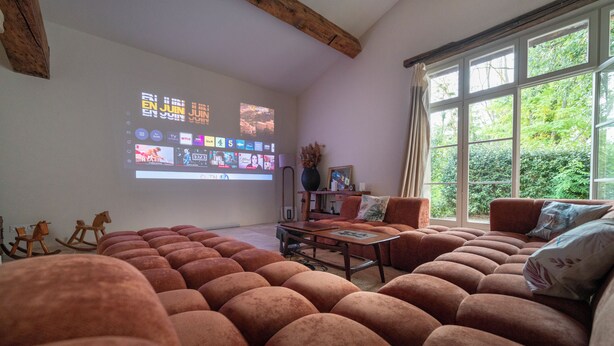 Un soggiorno con un grande schermo che mostra una guida TV, un divano, un tavolino da caffè e una finestra con vista su del verde.