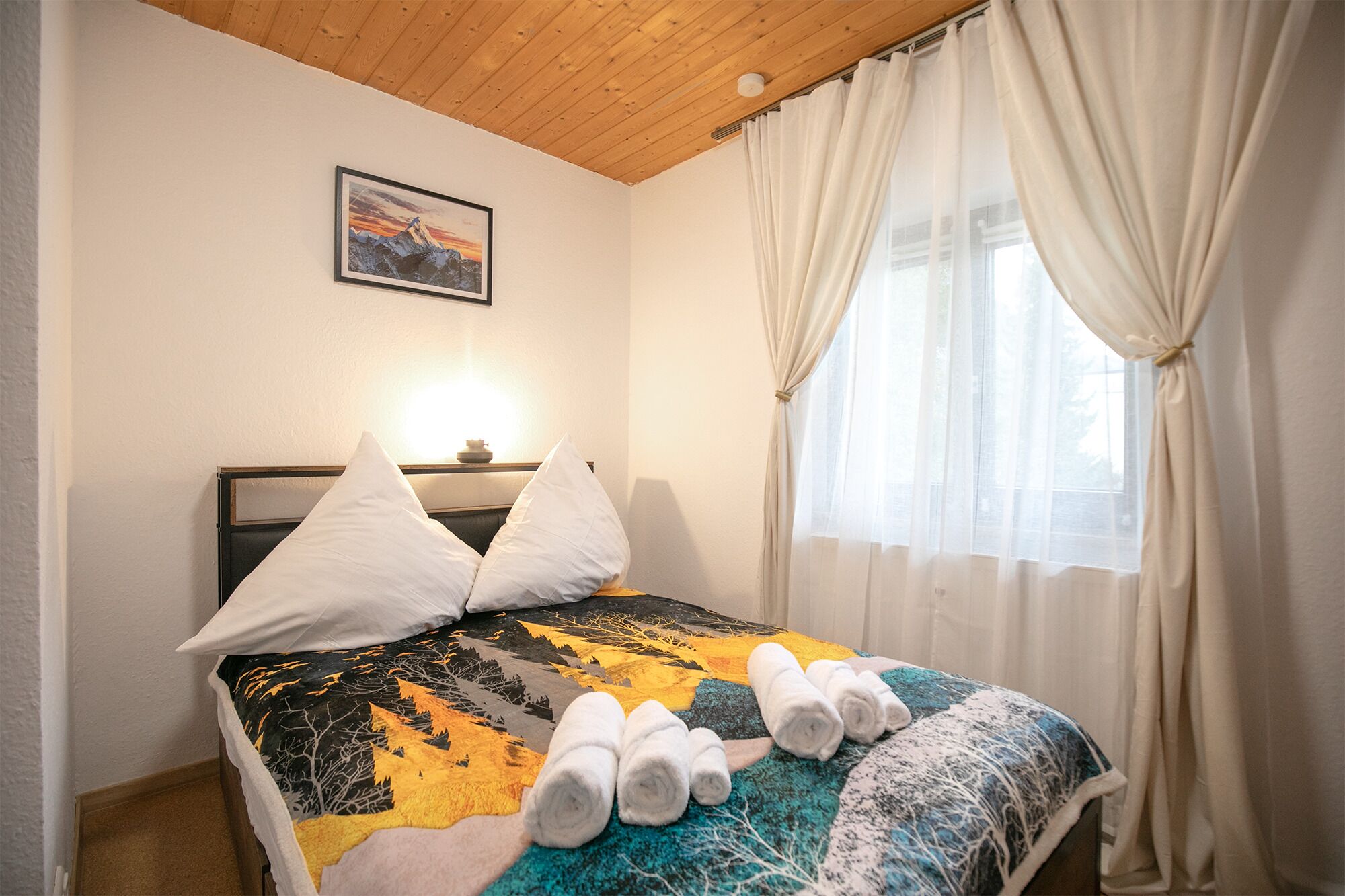 Een opgemaakt bed met een kleurrijke quilt, twee kussens en opgerolde handdoeken op het hoofdeinde.