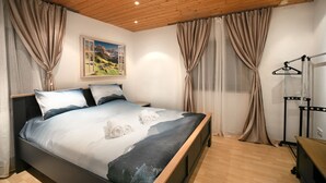 Une chambre avec un plafond en bois, un lit recouvert d’une courtepointe représentant un paysage de montagne, des rideaux beiges et un tableau représentant une scène de montagne.