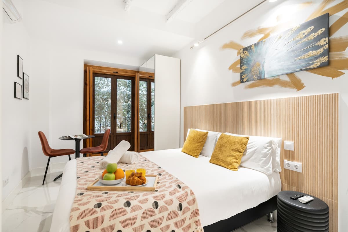 A modern hotel room with a large bed, a dining area with chairs, a table with breakfast, and a large window.