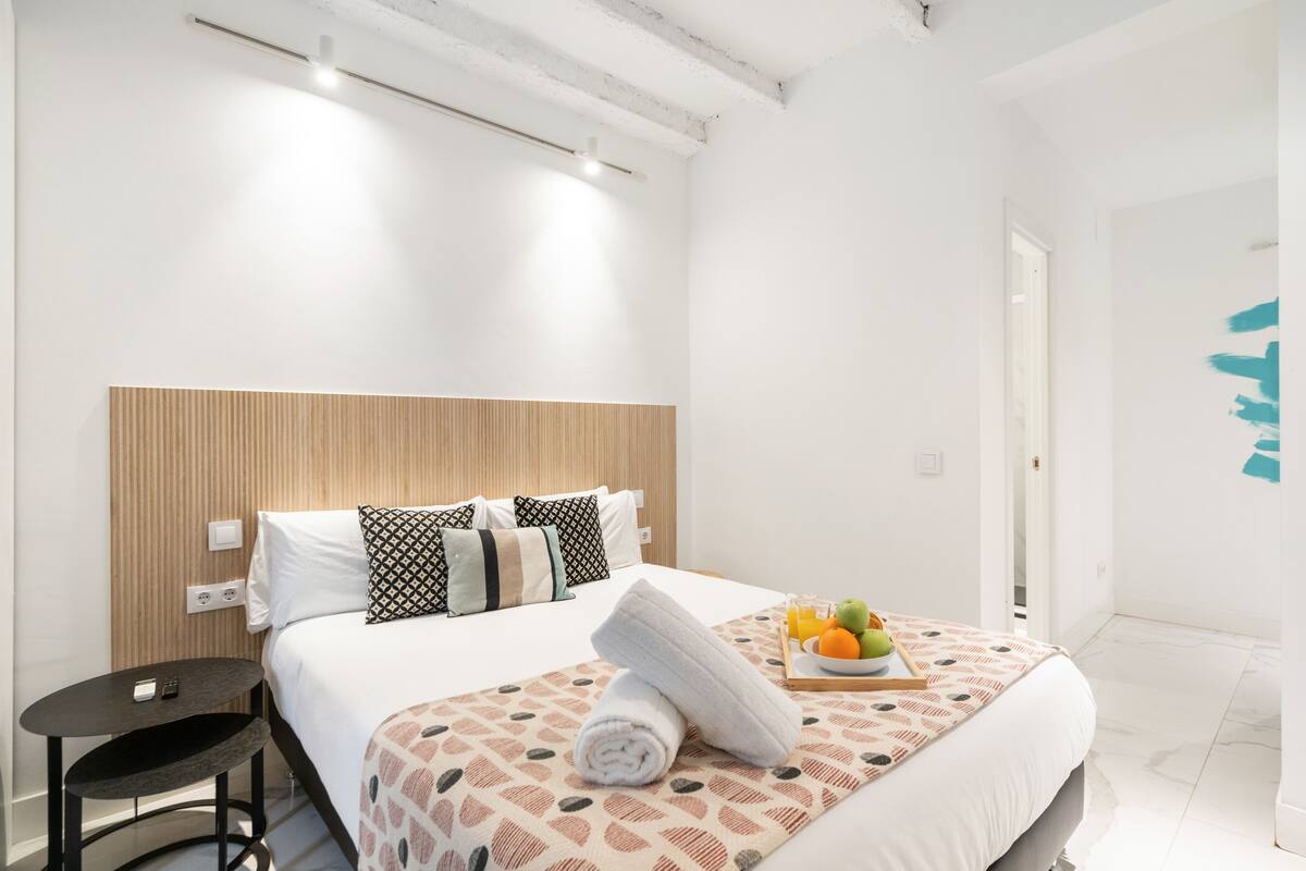 A hotel room with a bed, bedside table, and a tray with fruits.