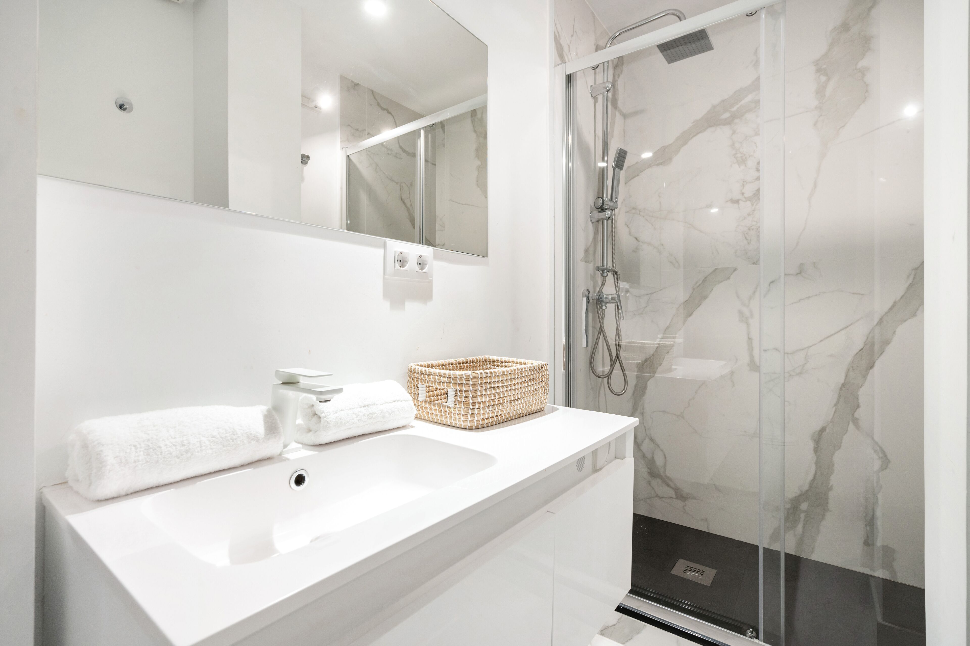 Un baño moderno con un lavabo blanco, un espejo, ducha con puerta de vidrio y pared de mármol.