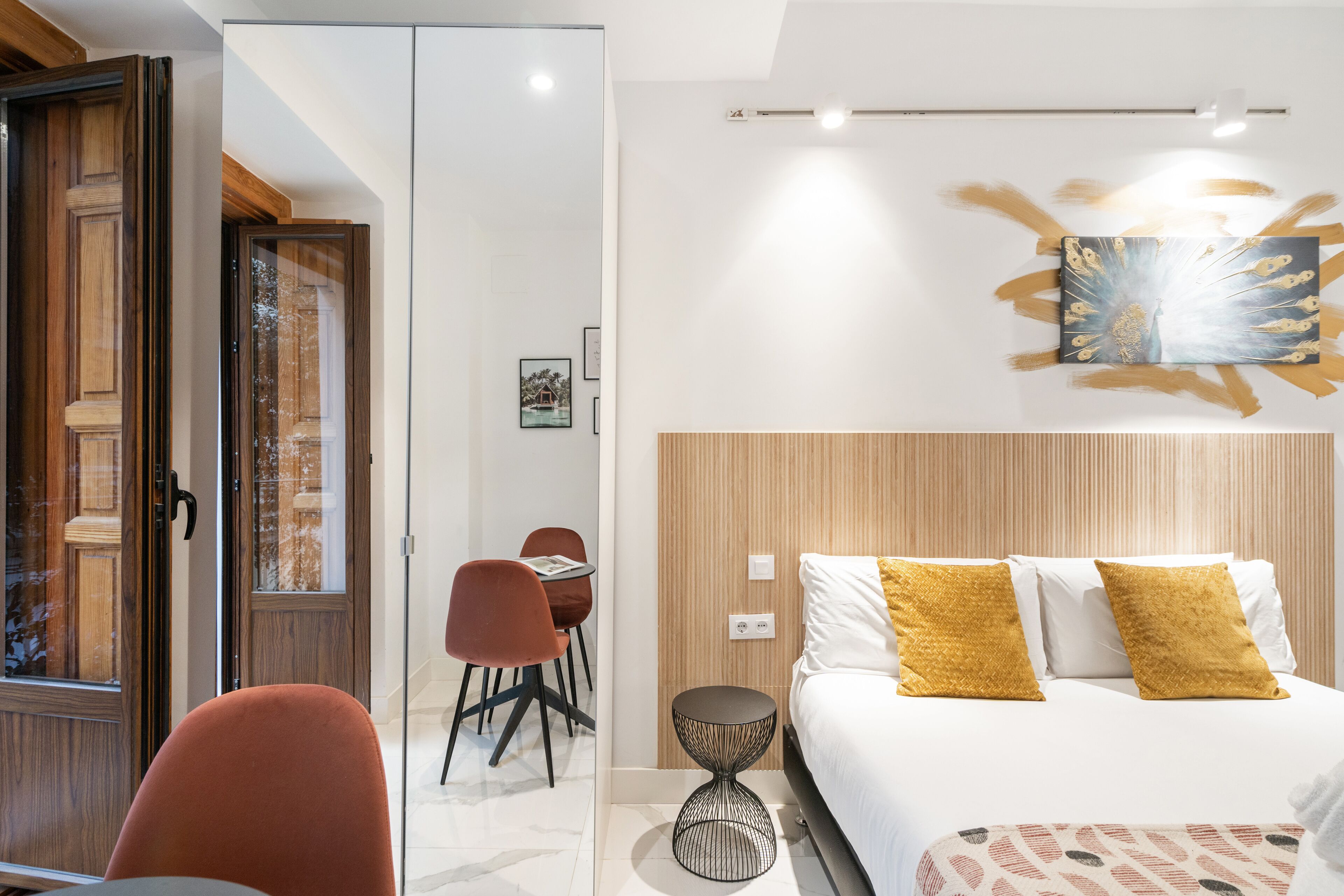 A modern hotel room with a bed, a round side table, a wooden headboard, and a dining area with chairs.