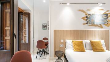 A modern hotel room with a bed, a round side table, a wooden headboard, and a dining area with chairs.