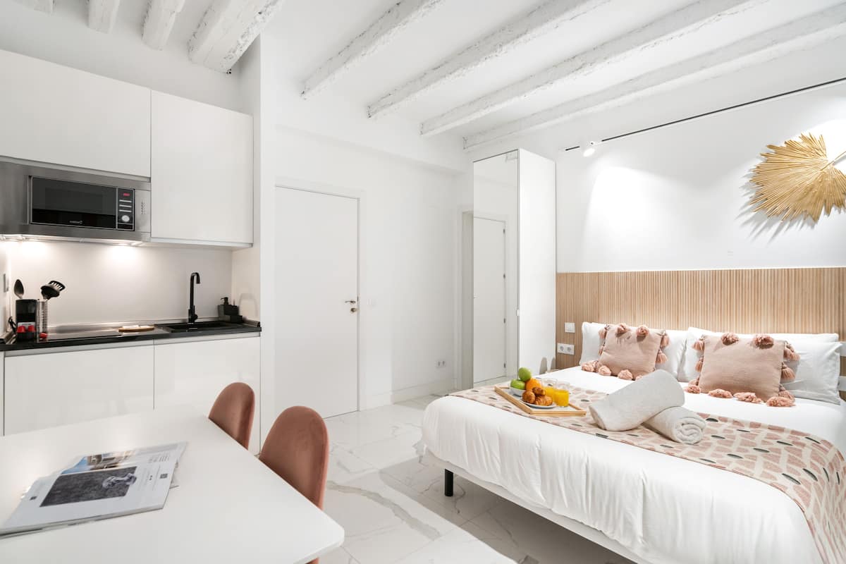 A modern hotel room with a kitchen area, a dining table, and a bed with a decorative headboard.