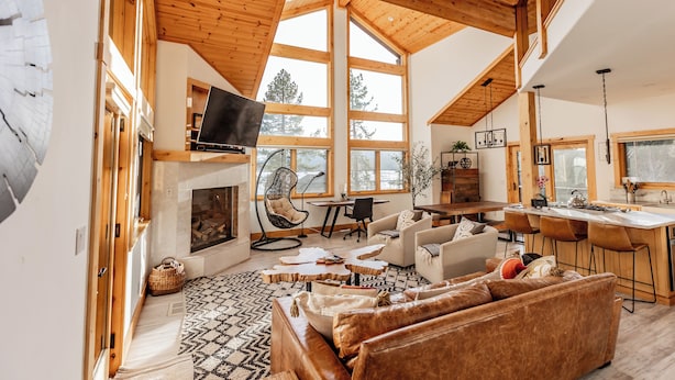 Amplia sala de estar con chimenea, techo de madera, ventanas grandes y zona de cocina.