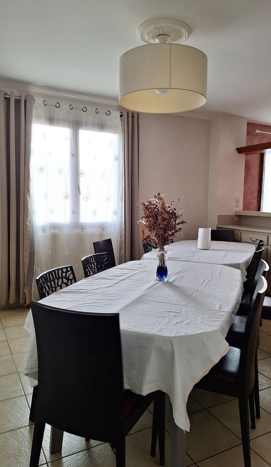 Een eethoek met een ronde tafel, bedekt met een witte tafelkleed, omringd door zwarte stoelen. Op de tafel staat een vaas met gedroogde bloemen.