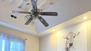 A bedroom with a bed, a ceiling fan, a wall-mounted light, a window with curtains, and a wall art of a cow. - Cowtown at Piney River Ranch; Charming Farm House in Houston, MO (Houston)