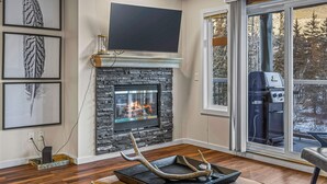 A living room with a stone fireplace, a flat-screen TV mounted above it, and a large window offering a view of the outdoors. - Welcoming 2 Bed/2 Ba-View-Pet-Gym-Parking-Wash/Dry (Canmore)