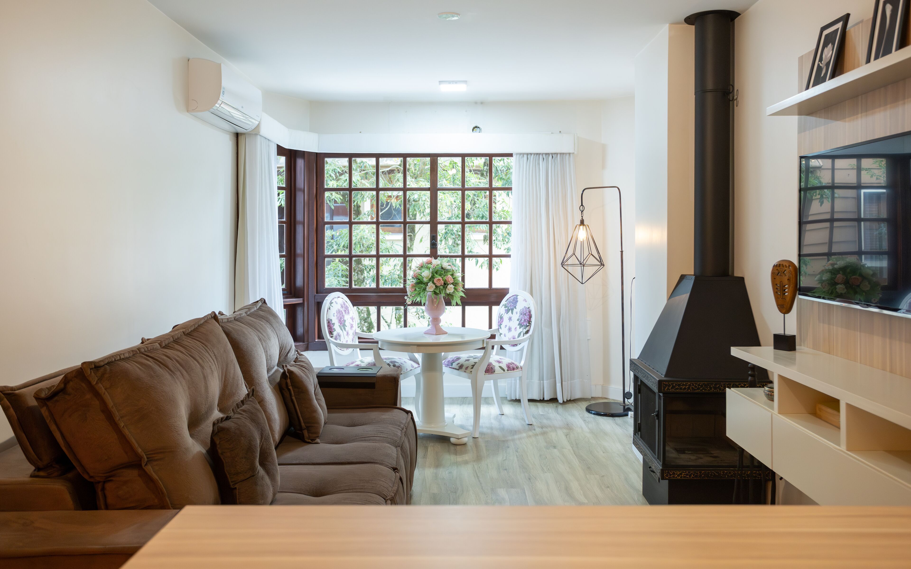 A cozy living room with a brown sofa, a round table with chairs, a fireplace, and a large window with curtains.