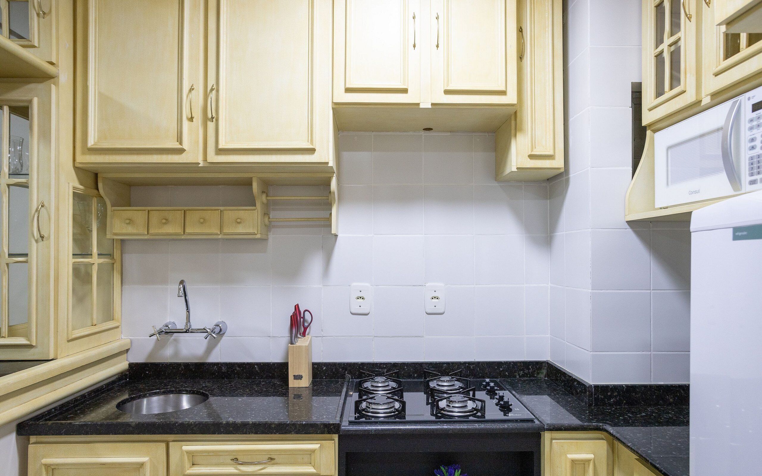 Una cocina con armarios de colores claros, encimera negra, pileta de acero inoxidable, estufa a gas y microondas.