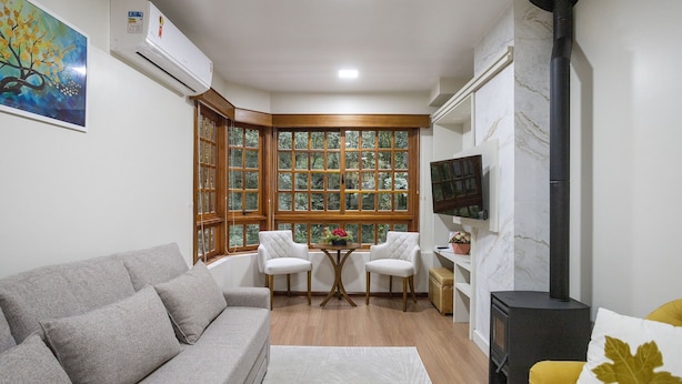 Ein Wohnzimmer mit grauem Sofa, einem runden Tisch mit Stühlen, einem an der Wand befestigten Fernseher und einem großen Fenster mit Blick auf Bäume.