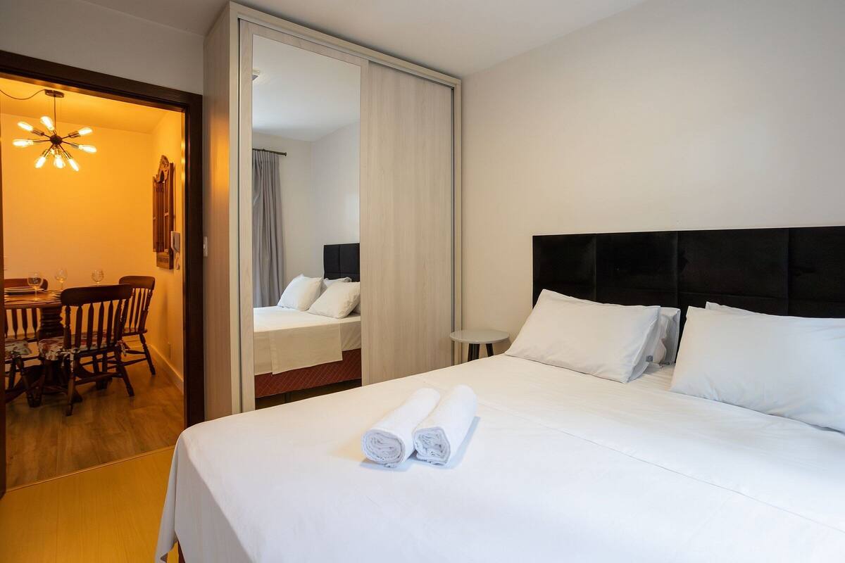 Una habitación de hotel con una cama grande, una mesita de noche pequeña, una silla y una zona de comedor con una mesa y sillas.