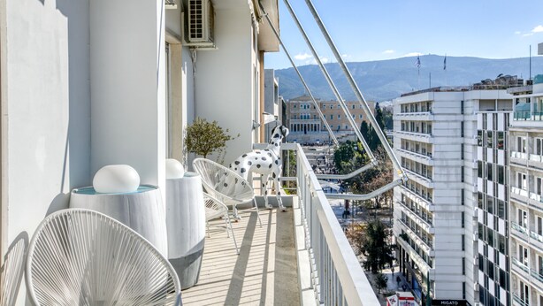 Un balcon avec des chaises blanches et une statue de chien, d’où l’on a une vue sur un paysage urbain avec des bâtiments et des montagnes.