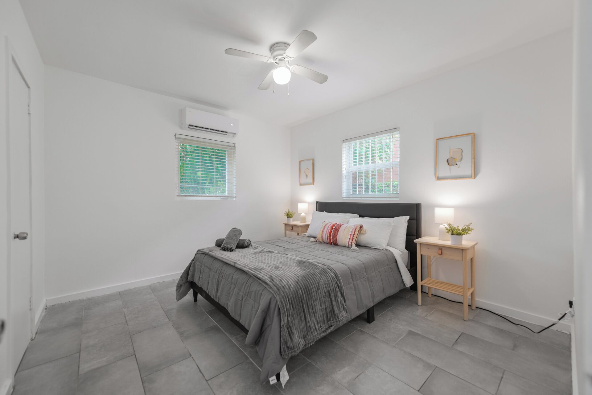 A bedroom with a bed, a ceiling fan, an air conditioner, two bedside tables with lamps, and a window with blinds.