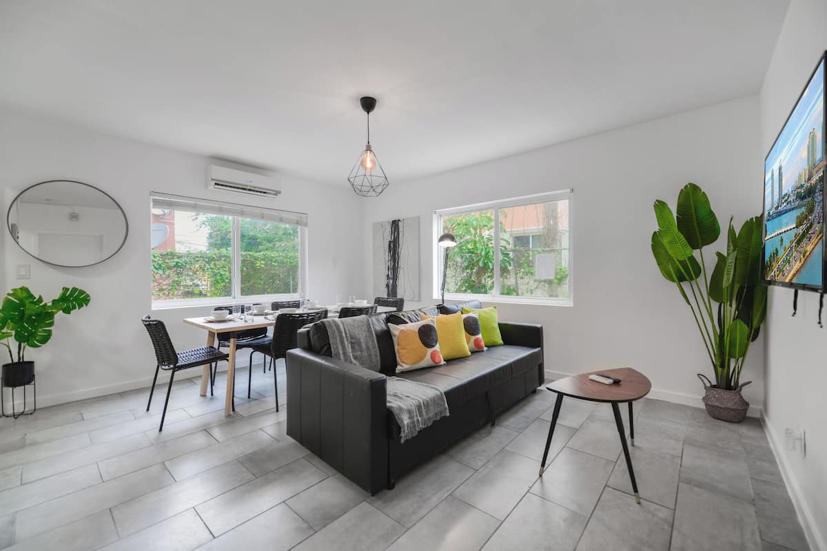 A modern living room with a black sofa, dining table, and chairs, large windows, and a potted plant.