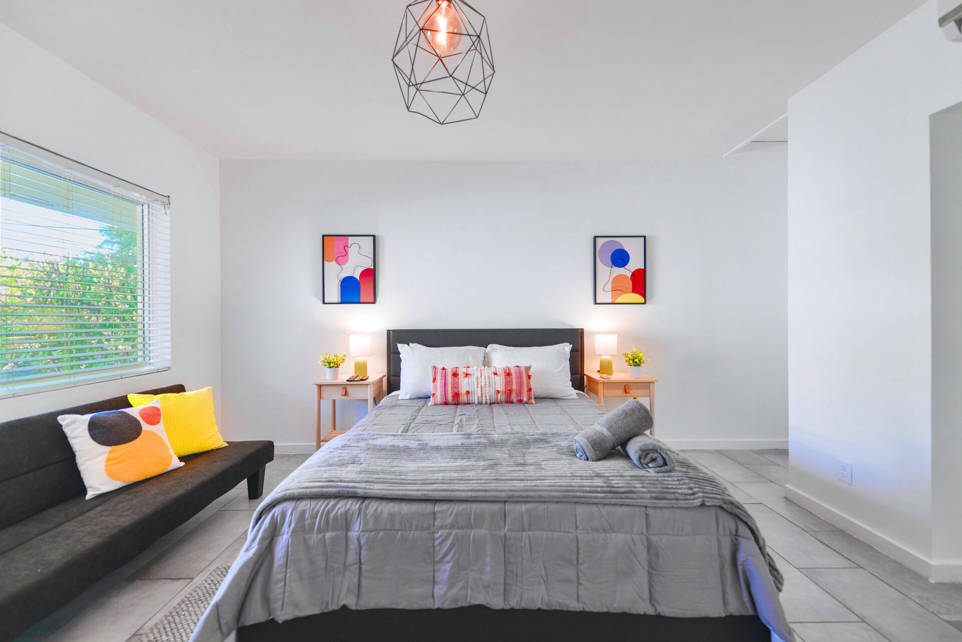 A modern bedroom with a large bed, two bedside tables with lamps, a black sofa with colorful cushions, and abstract wall art.