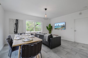 Un espace repas moderne avec une table en bois dressée pour quatre personnes, un canapé gris et une grande fenêtre donnant sur un paysage verdoyant.
