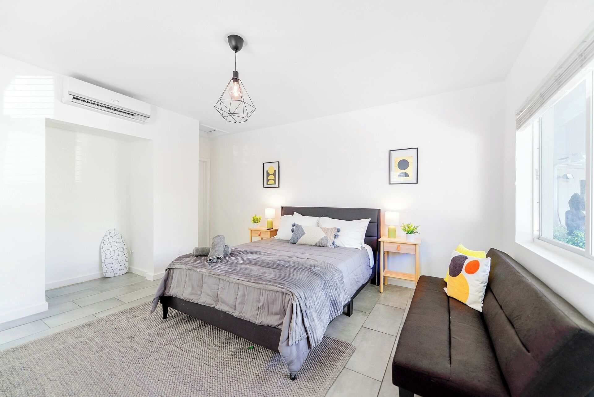 A modern bedroom with a bed, bedside tables, a window, and an air conditioning unit.