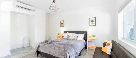 A modern bedroom with a bed, bedside tables, a window, and an air conditioning unit.
